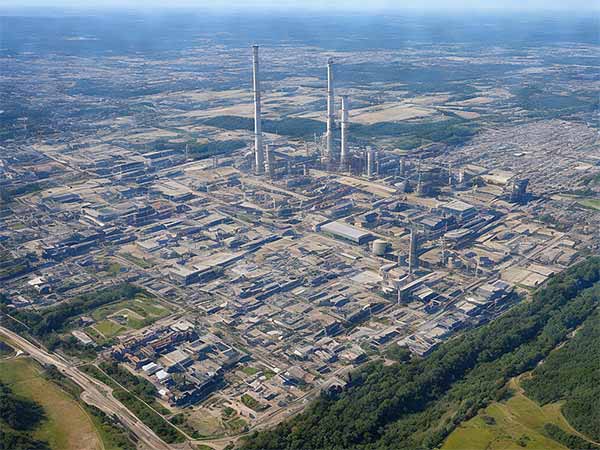 Luftaufnahme eines großen Industriekomplexes mit mehreren Gebäuden und hoch aufragenden Schornsteinen, umgeben von grünen Feldern.