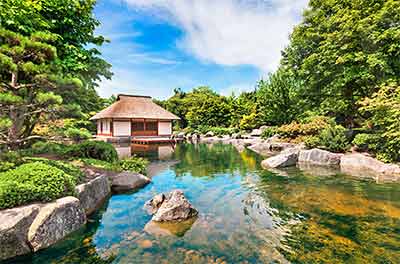 Ruhiger japanischer Garten mit einem Pavillon mit Strohdach an einem klaren Teich, umgeben von üppigem Grün und Felsen, unter einem strahlend blauen Himmel mit vereinzelten Wolken.