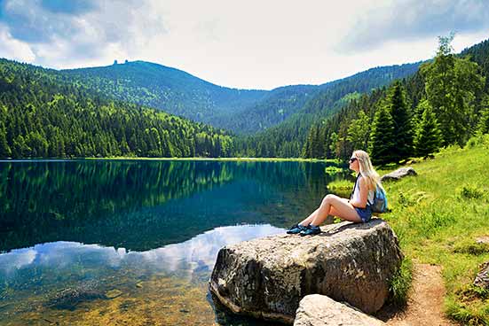 Eine Person mit Mütze und Rucksack sitzt auf einem großen Felsen neben einem ruhigen See, umgeben von bewaldeten Hügeln (Nationalpark Bayerischer Wald).