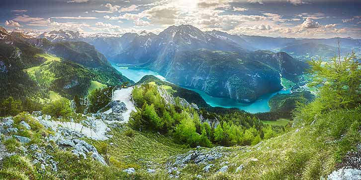 Ein Panoramablick auf eine Berglandschaft mit üppigem Grün, felsigen Gipfeln und einem türkisfarbenen Seen unter einem teilweise bewölkten Himmel.