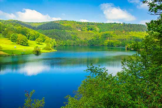 Ein ruhiger See, umgeben von üppig grünen Hügeln und Bäumen unter einem klaren blauen Himmel mit ein paar vereinzelten Wolken.