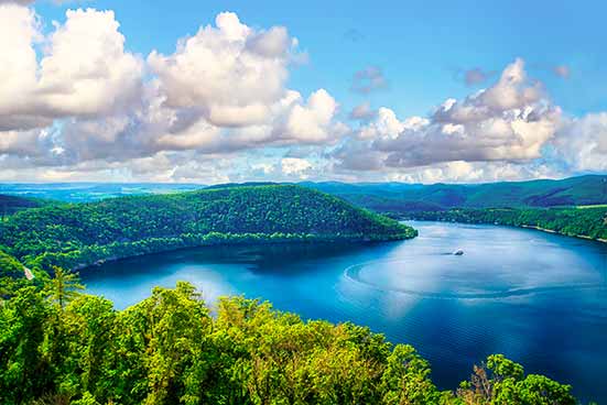 Eine malerische Aussicht auf einen ruhigen See, umgeben von üppigen grünen Hügeln unter einem blauen Himmel mit vereinzelten Wolken. Auf dem See ist ein Boot zu sehen.
