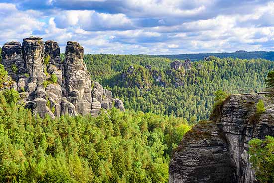 Unter einem bewölkten Himmel erheben sich Felsformationen über einem dichten Wald und zeigen eine natürliche Landschaft mit üppigem Grün und schroffen Klippen.