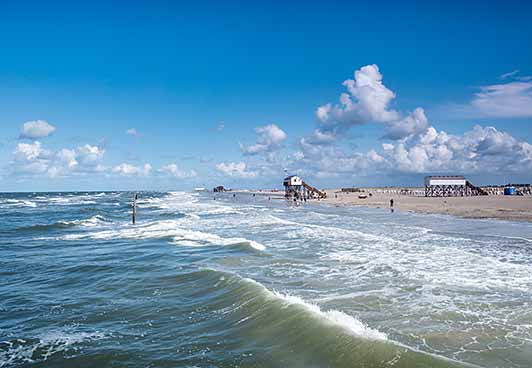 Ein Sandstrand mit sanften Wellen, blauem Himmel und weißen Wolken. Entlang der Küste sind Gebäude und Menschen zu sehen, darunter ein Gebäude auf Stelzen.